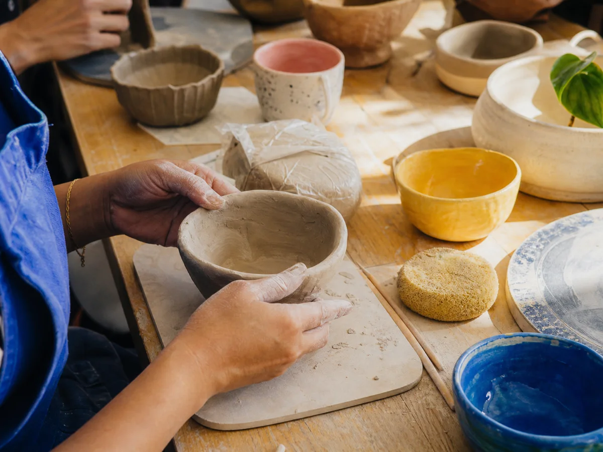 Hands shaping a clay bowl at a sunlit community pottery table